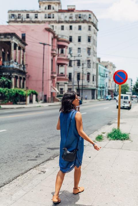 Woman in a blue dress with a black purse walking along a Havana sidewalk past faded pink apartment buildings