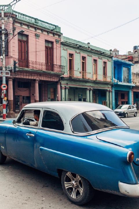 Man at the wheel of a classic blue car with a white roof passing pastel colonnaded buildings in Havana