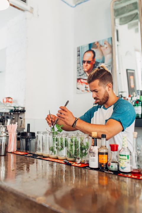 Bartender with a topknot muddling mint in a row of glass jars behind a Havana bar