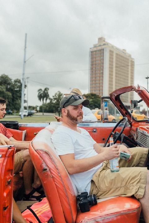 Bearded man in a cap and white t-shirt holding a canned drink in the front seat of a red convertible in Havana