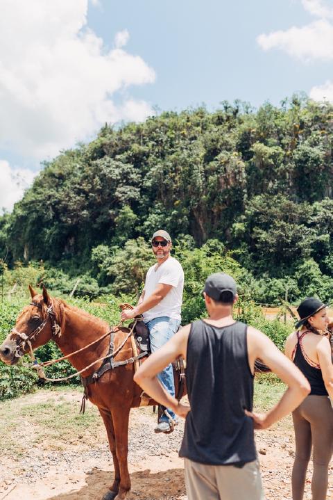 Man in a cap and white t-shirt on a chestnut horse below a jungle-covered hill in rural Cuba, others standing nearby