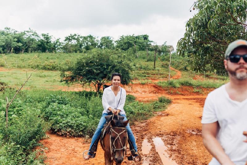 Woman in a white shirt riding a horse up a red-dirt trail through green Cuban countryside