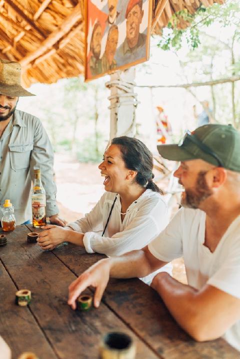 Travelers laughing over honey and rum at a wooden table under a thatched shelter in rural Cuba