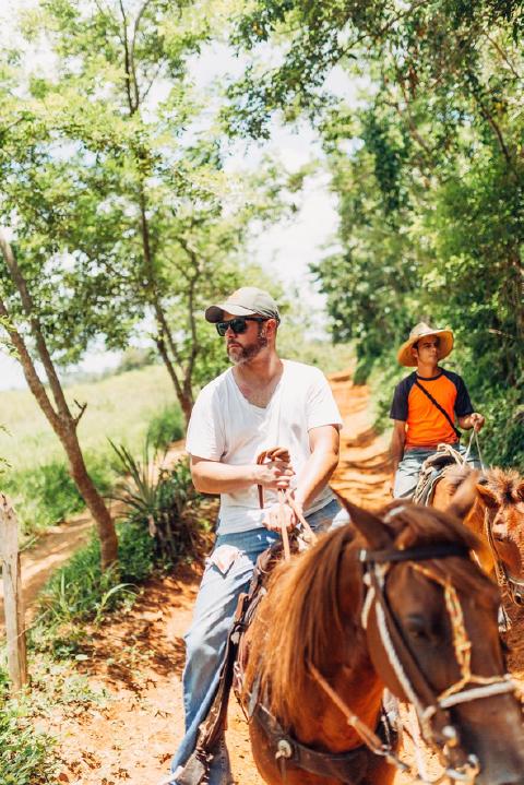 Two men on horseback riding a red-dirt trail between sunlit trees in the Cuban countryside