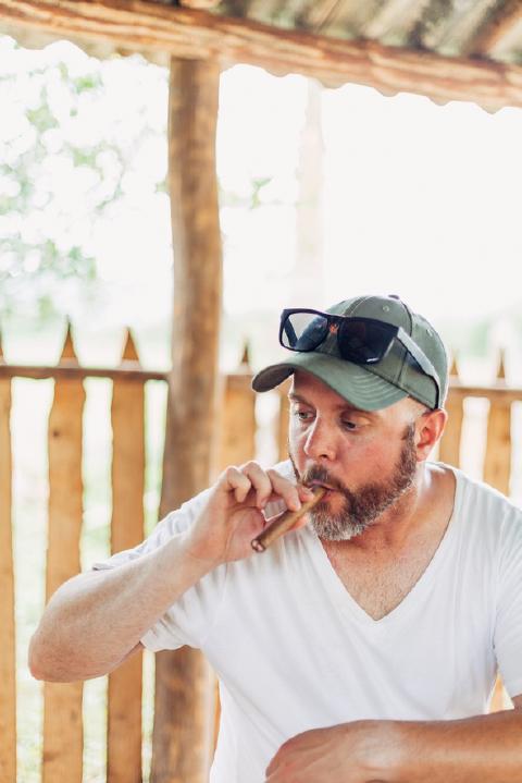 Bearded man in a green cap lighting a cigar beside a wooden picket fence under a rustic Cuban shelter