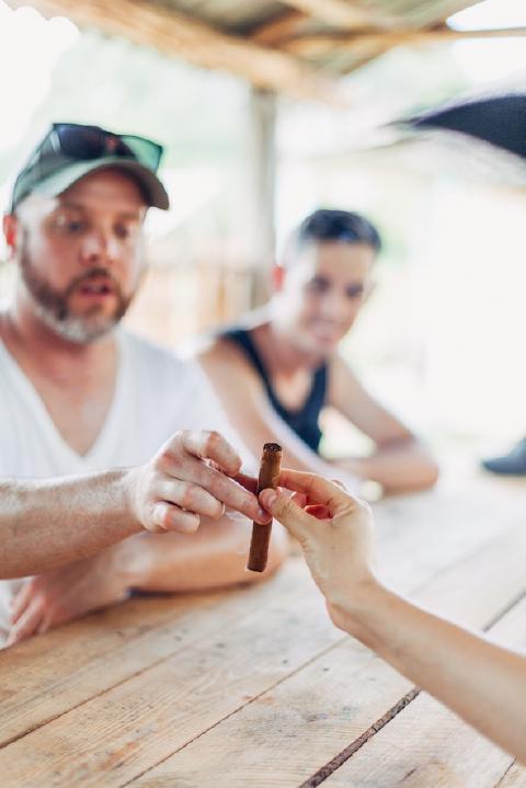 Hands passing a hand-rolled cigar across a wooden table under a thatched roof in rural Cuba