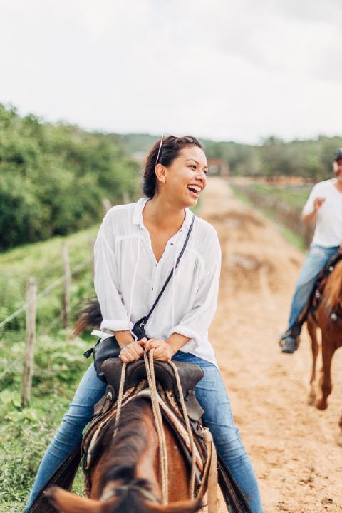 Laughing woman in a white shirt and jeans gripping her saddle on a dirt road in the Cuban countryside