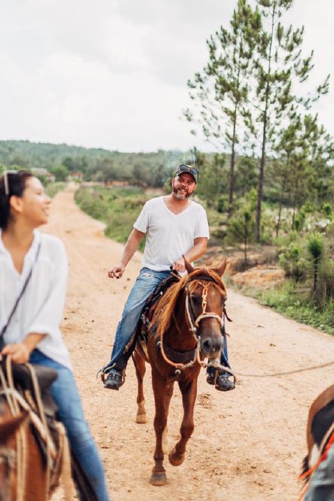 Man in a white t-shirt smoking a cigar on horseback along a red-dirt road lined with pines in Cuba