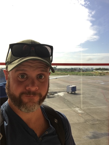 Wide-eyed selfie of a bearded man in a cap at an airport window with the tarmac behind him