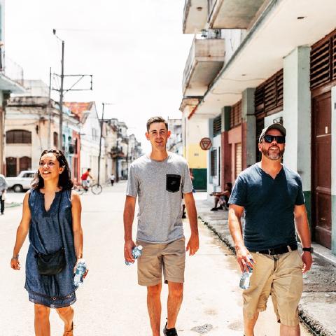 Three travelers carrying water bottles walking down a sunny Havana street of faded storefronts