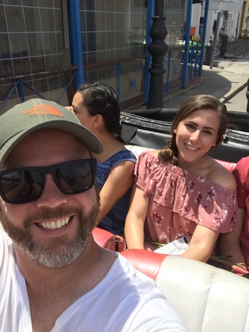 Selfie of a smiling man in a green cap riding in an open classic car with two women in the back seat