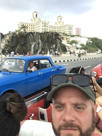 Selfie from an open car passing a blue vintage sedan, with the twin towers of Havana&rsquo;s Hotel Nacional behind