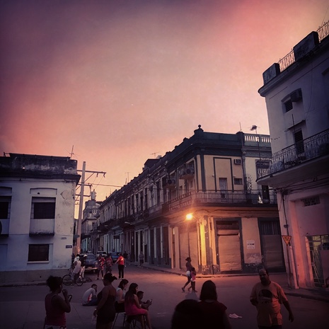 People gather on a Havana street corner at dusk under a pink sky, one streetlight glowing on the old buildings