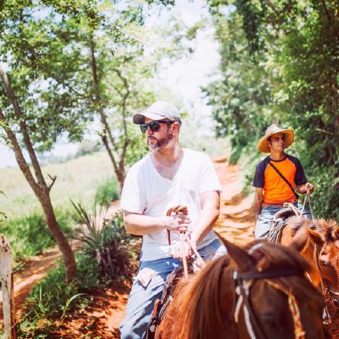 Two men ride horses down a red-dirt trail through green trees in Cuba, the lead rider in a cap and sunglasses