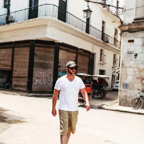 A man in a white t-shirt and cap crosses a sunlit Havana street corner, a bicycle taxi passing behind him