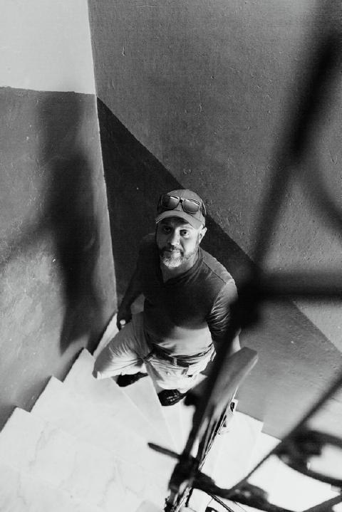 Black-and-white view down a stairwell in Cuba, a bearded man in a cap on the steps looking up at the camera