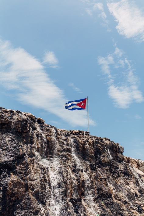 Cuban flag flying on a pole atop a rugged rock outcrop against a blue sky with wispy clouds