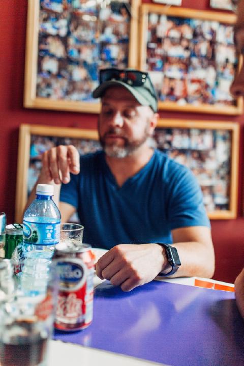 A bearded man in a cap sits at a cafe table in Cuba with soda cans and a water bottle, framed photo collages on the red wall