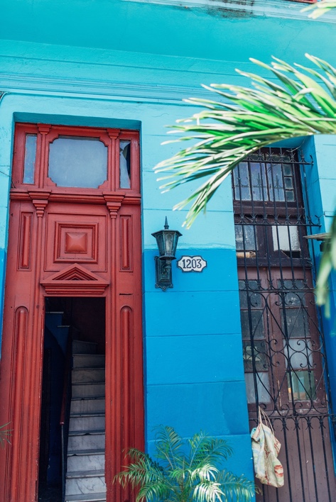 Bright turquoise house facade in Cuba with a red wooden doorway, wrought-iron window bars, and palm fronds overhead