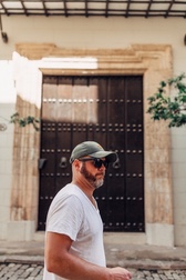 A man in a green cap and white t-shirt walks past a large carved wooden door on a stone building in Cuba