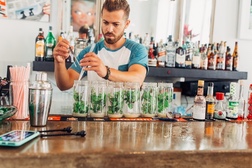 A bartender in Cuba muddles mint in a row of glasses for mojitos, liquor bottles lining the shelves behind him