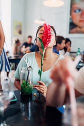 A woman at a busy Cuban cafe table holds a red tropical flower in front of her face, a mojito glass nearby