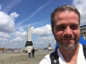 Selfie of a bearded man on a wide Brussels plaza, a tall white war memorial column rising behind him