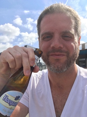 Close selfie of a smiling bearded man holding up a bottle of Hoegaarden beer under a partly cloudy Belgian sky
