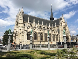 Gothic church with a slender spire beside a Brussels square ringed by columns topped with green statues