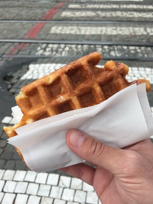 A hand holds a sugar-crusted Belgian waffle in a paper wrapper above cobblestones and tram tracks