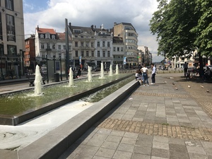 Long fountain pool with rows of small water jets along a Brussels street, people strolling past old buildings