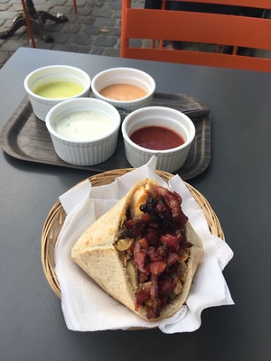 Stuffed pita topped with diced tomatoes in a basket, four ramekins of sauces on a tray at an outdoor table in Belgium