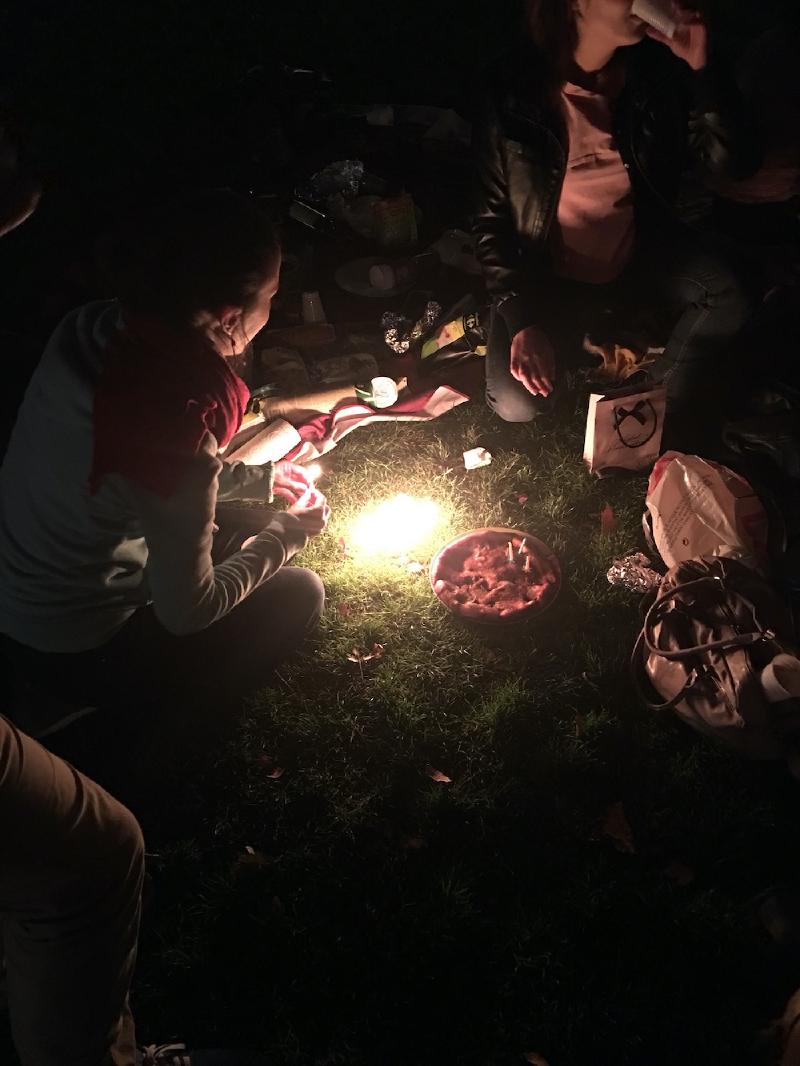 Friends sit on dark grass at a nighttime picnic in Paris, a candle lighting a dessert with candles at the center