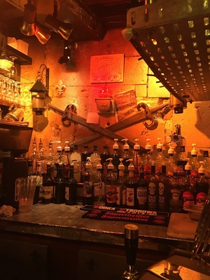 Dimly lit stone-walled bar in Caen with rows of vodka bottles, crossed swords, and lanterns glowing orange