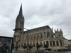 Gothic church with a tall pointed spire under heavy gray clouds in Caen, cars passing on the street below