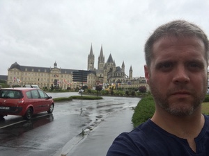 Selfie of a bearded man on a rainy street in Caen, the spires of a large abbey complex rising behind him