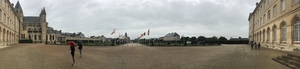 Panorama of a wide gravel esplanade with flagpoles between long stone buildings under a gray sky in Caen