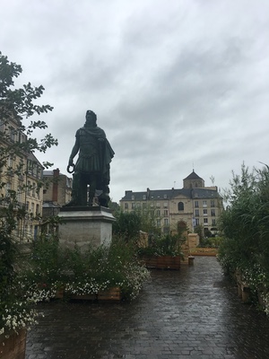 Bronze statue on a stone pedestal among flower planters on a rain-wet square in Caen, a domed church behind