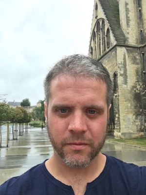 Close selfie of a bearded man on a wet street in Caen, the stone wall of a Gothic church rising beside him