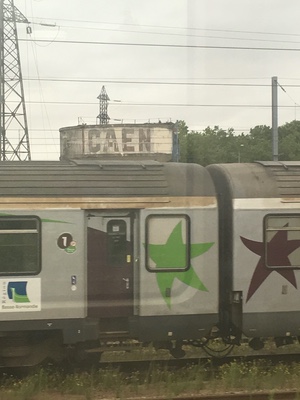 Silver regional train cars painted with green and maroon stars in a rail yard, CAEN lettered on a water tank behind