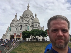 Selfie of a bearded man in front of the white domes of Sacre-Coeur basilica in Paris, crowds on the steps behind