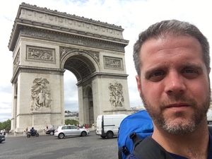Closer selfie of a bearded man beside the Arc de Triomphe, cars and vans circling its base