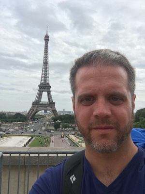 Selfie of a bearded man with the Eiffel Tower rising behind him under an overcast sky