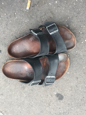 A pair of well-worn leather sandals with black buckled straps sitting on pavement