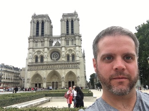 Selfie of a bearded man in front of the twin-towered facade of Notre-Dame cathedral in Paris