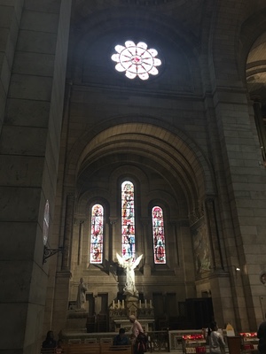 Dim stone chapel in Paris with a rose window above tall stained-glass panels and an angel statue at the altar