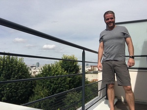 A smiling man in a t-shirt and shorts stands on a rooftop terrace railing with the Paris skyline behind