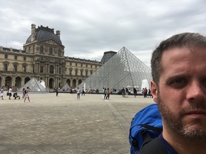 Selfie of a bearded man wearing a blue backpack in the Louvre courtyard, glass pyramid and palace wings behind