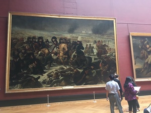 Visitors stand before a wall-sized painting of a cavalry battle scene in a red-walled Louvre gallery