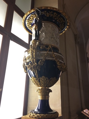 Tall cobalt-blue porcelain urn with ornate gilt handles and garlands beside a Louvre window
